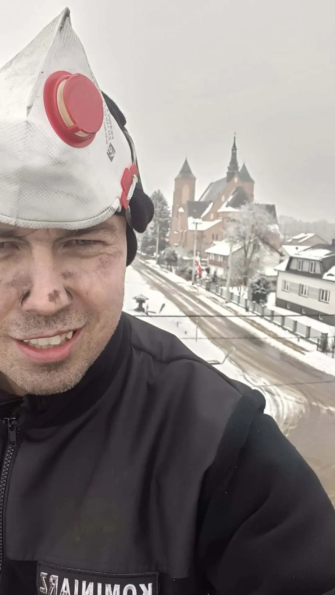 Piotr Szabłowski na dachu w masce przeciwpyłowej i bluzie z napisem KOMINIARZ